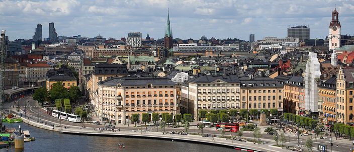 An elevated summer view over a curving stone quay and the tall ochre and pale-rendered blocks of Stockholm's old town, a dense roofline of green copper and tiled roofs behind it, a slender dark church spire and a clock tower rising above, water in the foreground.