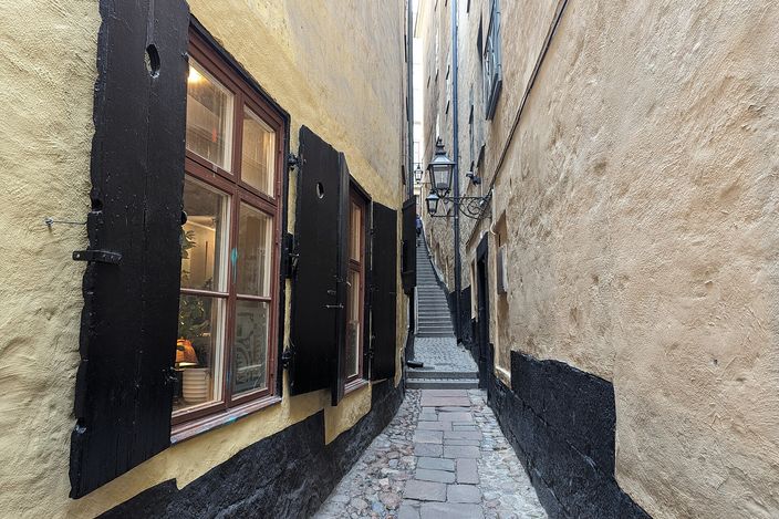 A very narrow alley in Stockholm's old town, barely a shoulder's width, running between an ochre wall with heavy black shutters and a bare rendered wall, its cobbled floor rising to a flight of steps under a wrought-iron lantern.