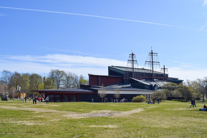 The dark timber-clad Vasa Museum on Djurgården seen across an open lawn, three tall replica ship's masts rising from its roof against a clear blue sky, small groups of visitors walking towards the entrance.