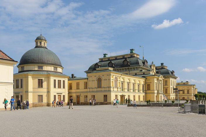 Drottningholm Palace in sunshine seen from its forecourt: a long pale-yellow baroque range with a black mansard roof and a pedimented centre, and beside it the palace chapel under a dark domed cupola, visitors crossing the gravel in front.