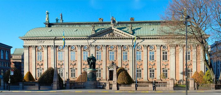 The long baroque facade of Riddarhuset in Stockholm: a pink-rendered palace with pale pilasters, a curved green copper roof and rooftop figures, a dark bronze statue on a plinth in front of it and clipped conical hedges along a low stone balustrade, under a clear blue sky.