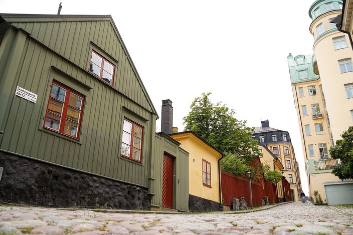 A cobbled lane on a Södermalm cliff-top: a green wooden cottage and a small ochre outbuilding on the left, a domed stone apartment block behind, greenery and pale render buildings down the slope.