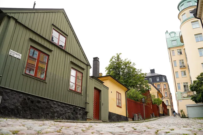 A cobbled lane on a Södermalm cliff-top: a green wooden cottage and a small ochre outbuilding on the left, a domed stone apartment block behind, greenery and pale render buildings down the slope.