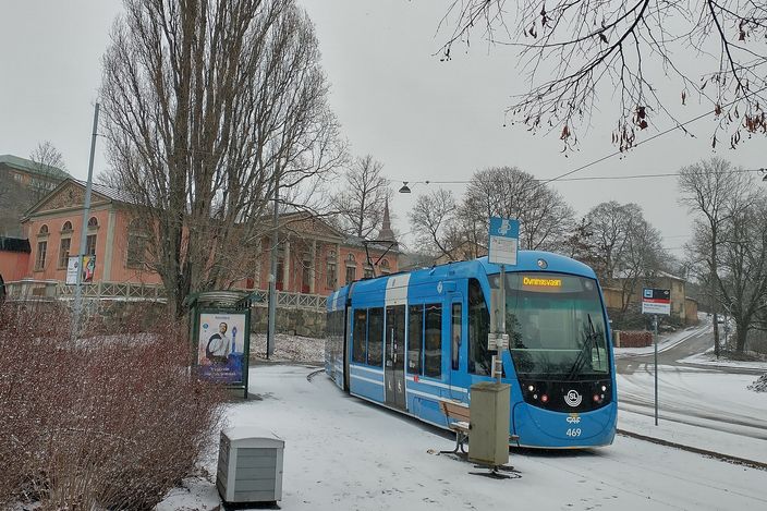 A blue SL tram bound for Djurgården halted at a stop near Skansen, bare winter trees and a light dusting of snow on the ground, a pale historic building behind.