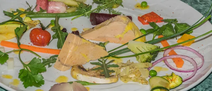 Slices of pale pink foie gras terrine on a white plate among small spring vegetables and herbs