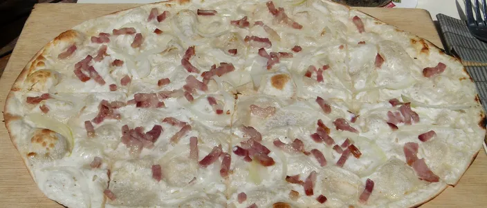 A thin, oblong tarte flambée on a wooden board, its white cream base scattered with onion and small strips of bacon