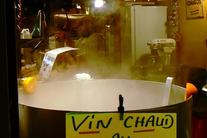 Steam rising from a large pot of mulled wine with halved lemons and oranges at its rim, a hand-lettered VIN CHAUD sign on the front and a stallholder working behind it