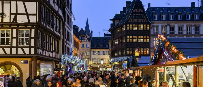 Crowds between lit wooden market stalls on Place de la Cathédrale at dusk, the timber-framed Kammerzell house and the old town's rooftops behind