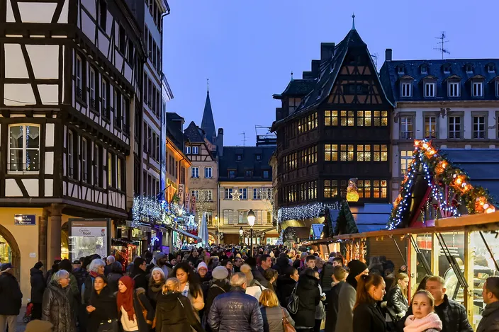 Crowds between lit wooden market stalls on Place de la Cathédrale at dusk, the timber-framed Kammerzell house and the old town's rooftops behind
