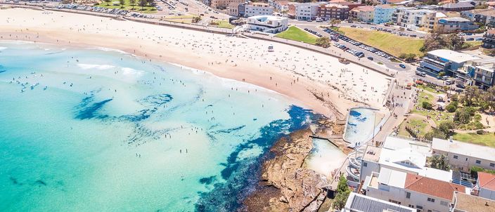 An aerial view of Bondi Beach's full sandy crescent packed with beachgoers, turquoise surf breaking along the shore, and the geometric white-and-turquoise Icebergs saltwater pool built into the rocks at the beach's southern end, with Sydney's inner suburbs on the horizon.