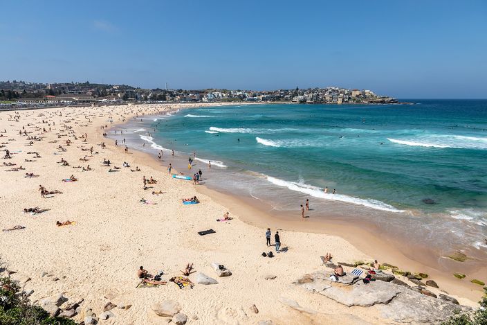 A crowded Bondi Beach seen from an elevated clifftop vantage: pale sand thronged with sunbathers, turquoise water breaking into white surf with swimmers and surfers scattered through it, and the shoreline curving away to the North Bondi headland and apartment blocks in the distance.