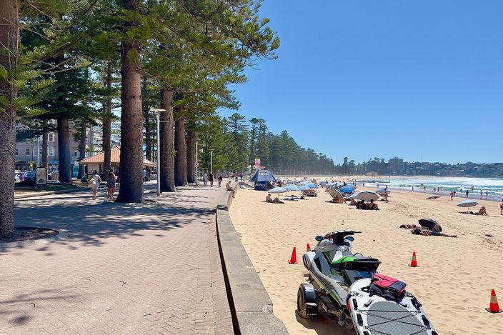 The Manly Beach promenade lined with tall Norfolk Island pines on the left, open sand and breaking surf on the right, with two jet-skis parked on the sand in the foreground and beachgoers under umbrellas further along.