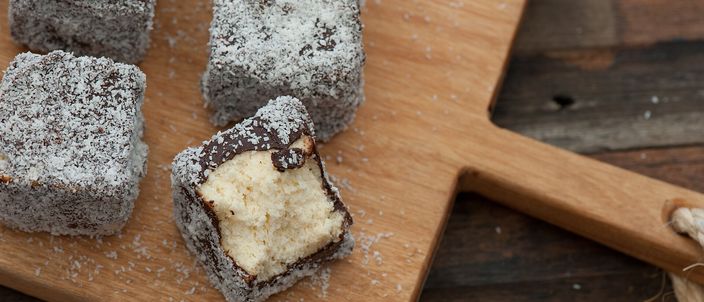 Squares of coconut-coated lamington on a wooden board, one cut open to show the pale sponge inside