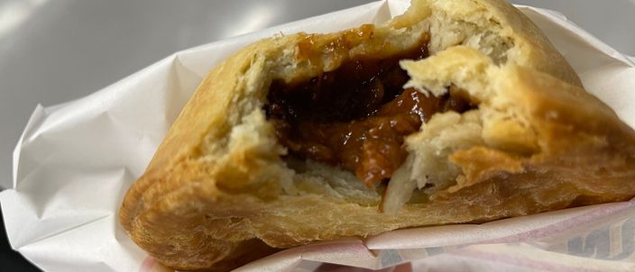 A hand holding an individual meat pie torn open in its paper bag, dark chunky beef filling showing