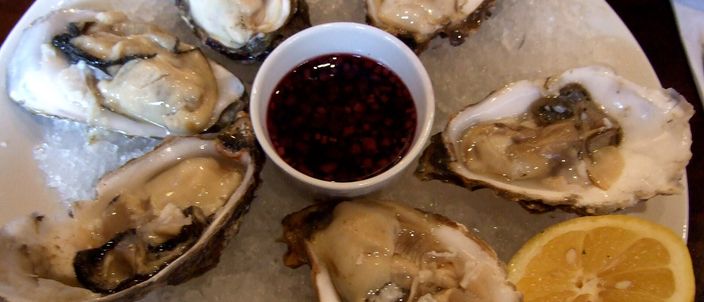 Six opened rock oysters on crushed ice around a small bowl of red mignonette dressing, with a lemon half