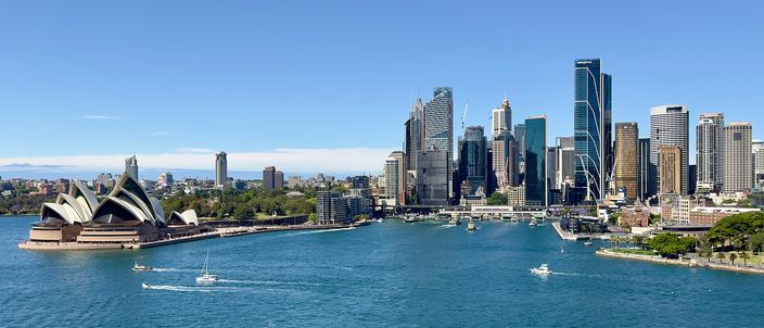 The white sails of the Sydney Opera House at left and the glass towers of the CBD at right, seen across the deep-blue harbour with small boats crossing, the arch of the Harbour Bridge just visible far right under a clear sky