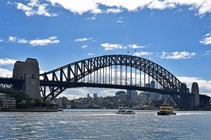 The steel arch of the Sydney Harbour Bridge seen across the harbour from water level, a granite pylon at each end, small ferries crossing beneath it and the low north-shore suburbs behind, under a bright sky with scattered cloud.