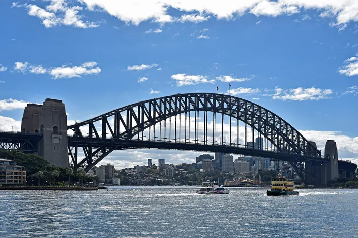 The steel arch of the Sydney Harbour Bridge seen across the harbor from water level, a granite pylon at each end, small ferries crossing beneath it and the low north-shore suburbs behind, under a bright sky with scattered cloud.