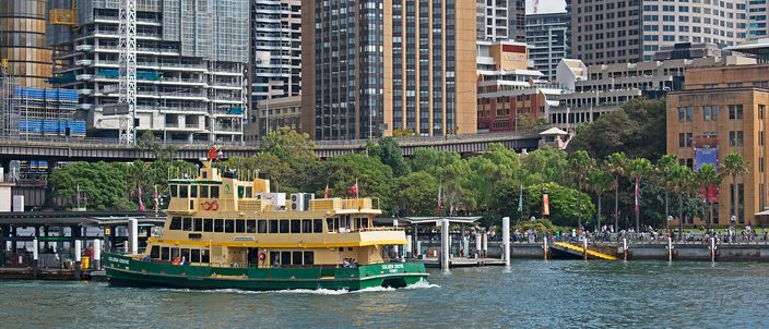 A green-and-yellow Sydney ferry crossing the water at Circular Quay, the wharf canopies and the elevated railway viaduct behind it and the glass towers of the central business district rising above.