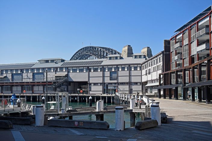 The Walsh Bay finger wharves seen from the water: long timber and corrugated-iron cargo sheds on piles running out into the harbour, a dark brick warehouse block at right, mooring posts and pontoons in the foreground, and the steel arch of the Harbour Bridge rising behind them.