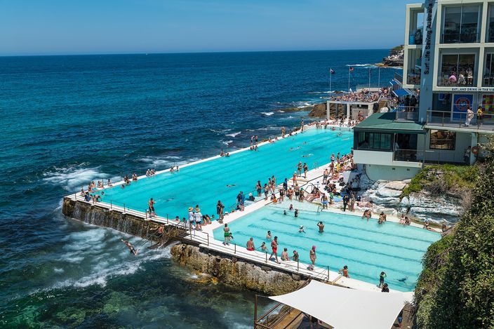 The Bondi Icebergs ocean pool from above: a long turquoise lap pool and a smaller shallow pool built onto a rock shelf at the end of the beach, crowded with swimmers, waves breaking white against the outer wall, and the club building at right.