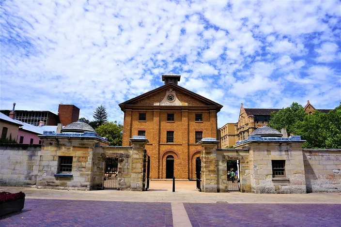 Hyde Park Barracks: a plain three-story Georgian brick building with a pedimented center and a small clock, seen head-on through the opening in its low sandstone perimeter wall, iron gates to either side and a paved forecourt in front under a mackerel sky.