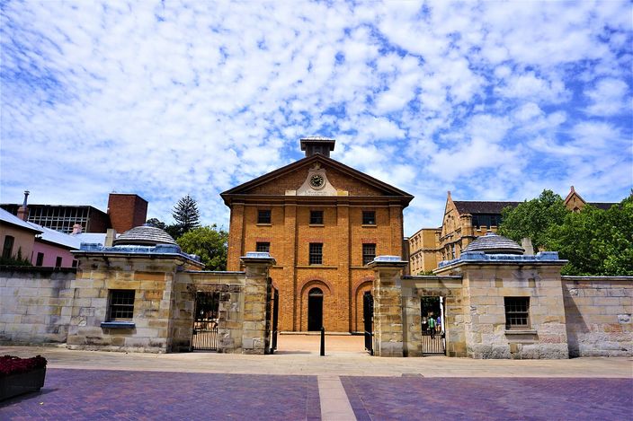 Hyde Park Barracks: a plain three-storey Georgian brick building with a pedimented centre and a small clock, seen head-on through the opening in its low sandstone perimeter wall, iron gates to either side and a paved forecourt in front under a mackerel sky.