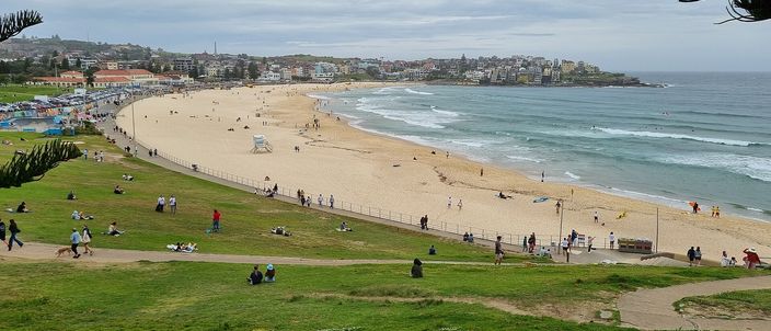 Bondi Beach seen from the grass of Bondi Park: a wide crescent of pale sand curving north to a low headland, white surf rolling in along its whole length, the low buildings of the suburb behind and people scattered across the lawn in the foreground under a grey sky.