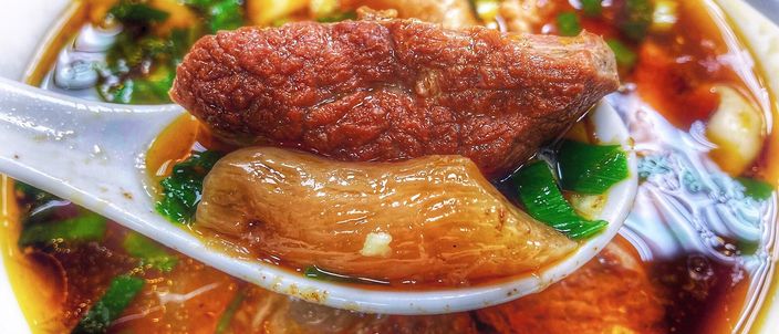 A bowl of Taiwanese red-braised beef noodle soup: wheat noodles and pieces of stewed beef in a dark, glossy broth, topped with greens.
