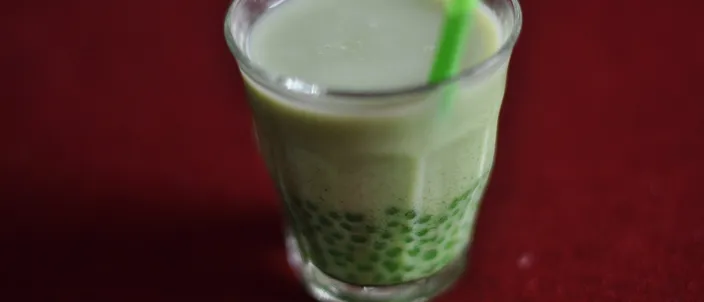 A clear glass of bubble tea: pale green matcha milk over a thick layer of dark tapioca pearls, with a straw standing in it.