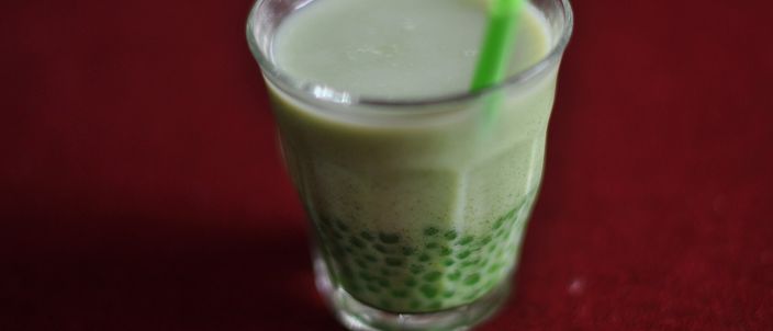 A clear glass of bubble tea: pale green matcha milk over a thick layer of dark tapioca pearls, with a straw standing in it.