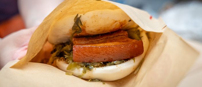 A gua bao held in paper: a soft folded white bun clamped around a slab of red-braised pork belly, with pickled greens and a dusting of peanut powder.