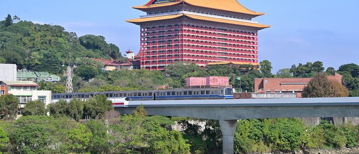 The Grand Hotel Taipei, a vast red pagoda-style building with a golden roof, rising on a wooded hillside above the Keelung River, with an MRT train crossing a concrete viaduct in the foreground under a clear blue sky