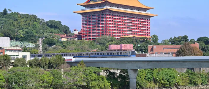 The Grand Hotel Taipei, a vast red pagoda-style building with a golden roof, rising on a wooded hillside above the Keelung River, with an MRT train crossing a concrete viaduct in the foreground under a clear blue sky