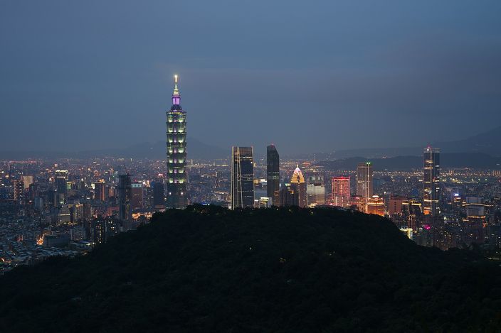 A city skyline at dusk seen from a hillside, with a tall tapering tower lit pale green standing above a spread of buildings, a dark wooded hill in the foreground and mountains behind.