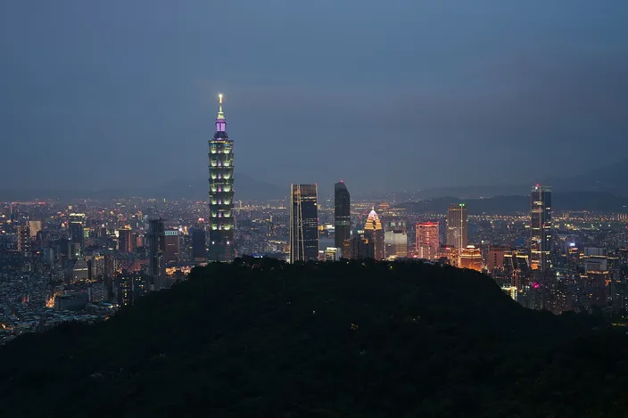 A city skyline at dusk seen from a hillside, with a tall tapering tower lit pale green standing above a spread of buildings, a dark wooded hill in the foreground and mountains behind.