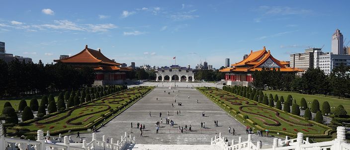 A vast paved square seen from above, with clipped hedges and topiary along both sides, a tall white gateway at the far end, and two large halls with orange tiled roofs facing each other across it under a blue sky.