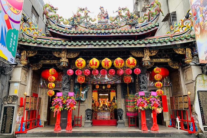 The front of a Chinese temple with a heavily carved and gilded facade, rows of red paper lanterns hung under the eaves, coloured ceramic dragons and figures along the roof ridge, and two tall flower arrangements flanking the central doorway.
