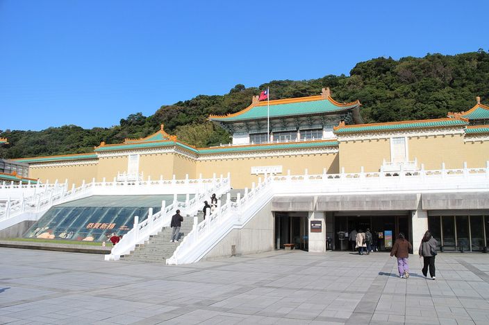 A long low museum building with pale yellow walls and green-and-gold tiled roofs standing against a wooded hillside, approached by wide white balustraded staircases across an empty stone forecourt, a flag on a pole above the central hall.