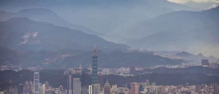 A dense city of towers filling a wide basin under ridge behind ridge of hazy blue-grey mountains, with one very tall tapered skyscraper rising far above everything else near the centre.