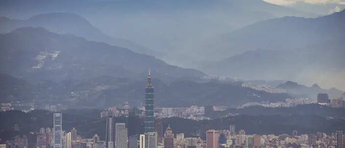 A dense city of towers filling a wide basin under ridge behind ridge of hazy blue-gray mountains, with one very tall tapered skyscraper rising far above everything else near the center.