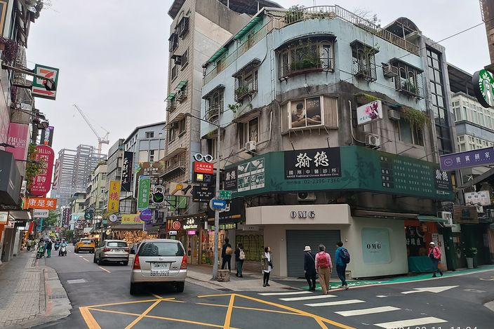 A Taipei street corner in Da'an: a plant-covered corner building with green shop signs, a 7-Eleven, low-rise shops, a zebra crossing and a few pedestrians under a pale sky.