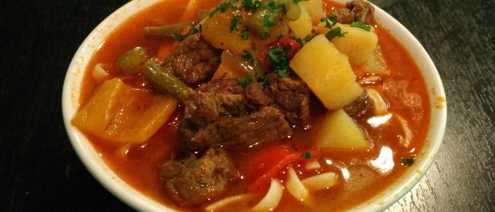 A bowl of soup lagman: reddish broth with chunks of stewed meat, potato and peppers over thick noodles, scattered with fresh herbs.