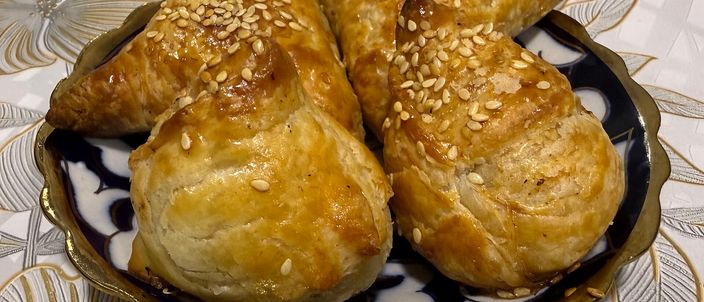 Three plump golden samsa, sealed into triangles and scattered with sesame seeds, on a decorative plate.