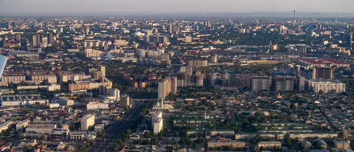 Aerial view over Tashkent at golden hour: a flat, spread-out city of low residential blocks, wide boulevards and green parks stretching to a hazy horizon with a distant tower and mountains beyond.