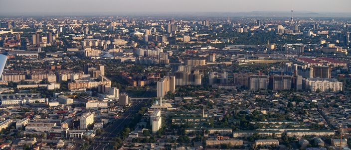 Aerial view over Tashkent at golden hour: a flat, spread-out city of low residential blocks, wide boulevards and green parks stretching to a hazy horizon with a distant tower and mountains beyond.