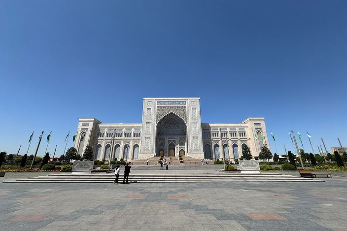 The Center for Islamic Civilization in Tashkent seen head-on across an empty paved forecourt: a broad white building with a tall pointed-arch portal at its centre, arcaded wings either side, rows of flagpoles flanking the approach and a few small figures on the steps, under a clear blue sky.