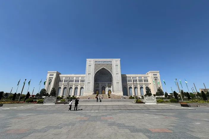 The Center for Islamic Civilization in Tashkent seen head-on across an empty paved forecourt: a broad white building with a tall pointed-arch portal at its center, arcaded wings either side, rows of flagpoles flanking the approach and a few small figures on the steps, under a clear blue sky.