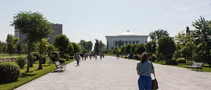 A wide paved pedestrian promenade through a park in Amir Timur Square, lined with trees, clipped topiary and benches, people walking away from the camera towards a dark equestrian statue with a large white domed building beyond under a hazy sky.
