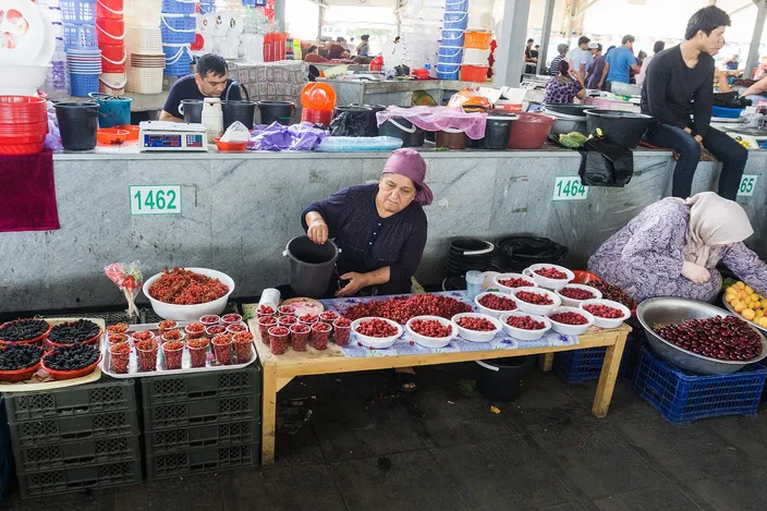 A stallholder in a headscarf sitting behind a trestle table at Chorsu Bazaar banked with bowls of cherries, redcurrants and dark berries, plastic crates stacked at her feet and other traders working along the concrete counter behind her under the market roof.